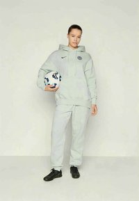 Junge Person in einem hellgrauen Kapuzenpullover und Jogginghosen, die einen Fußballs hält, mit schwarzen Fußballschuhen, steht vor einem schlichten hellen Hintergrund.