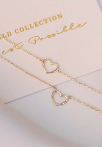 Gouden hartvormige ketting met een delicate ketting, voorzien van twee open hartpendants. Geplaatst op een lichte kaart met gouden tekst.
