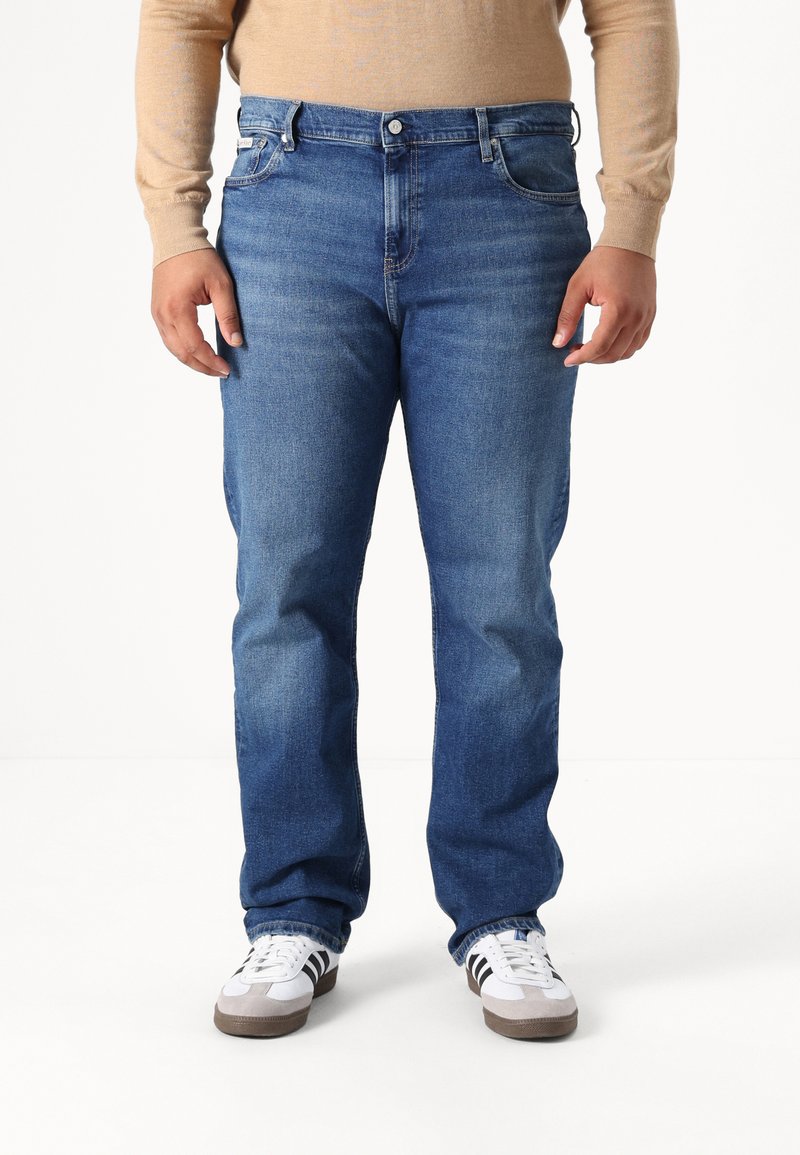 Jeans de mezclilla azul con un corte recto, diseño de cinco bolsillos y un ligero desgaste. Combinados con zapatillas blancas con franjas negras.
