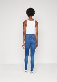 Jeans ajustados de color azul hechos de denim, con una cintura alta, dos bolsillos traseros y un pequeño parche de logo en la parte superior de la espalda. Se lleva una camiseta sin mangas blanca.