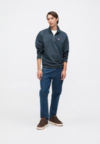 Pullover cinza escuro com colarinho de meia-zíper e um pequeno logotipo, combinado com jeans de denim azul e ténis castanhos com detalhes em creme.