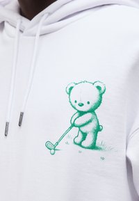 Λευκή μπλούζα με κουκούλα με πράσινο αρκουδάκι να παίζει γκολφ, κρατώντας ένα κλαμπ έτοιμο να χτυπήσει μια μπάλα γκολφ στο γρασίδι.