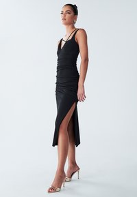 Black ruched dress with thin straps, V-neck, and high side slit; smooth fabric with a fitted design. Model wears gold heeled sandals.