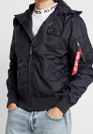 Mand iført en sort bomberjakke med lynlås med en rød "Remove Before Flight" tag på hylsterlommen og lyseblå jeans.