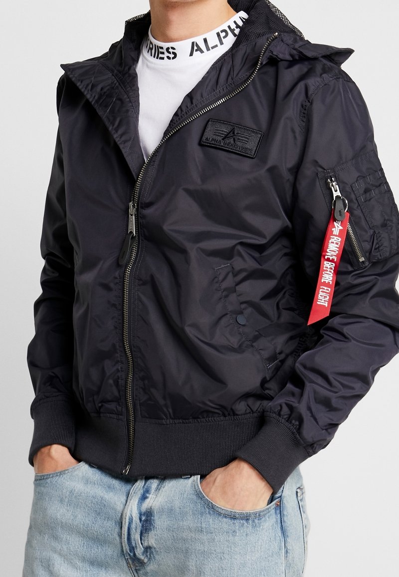 Homme portant une veste bomber noire zippée avec une étiquette rouge "Remove Before Flight" sur la poche de la manche et un jean bleu clair.