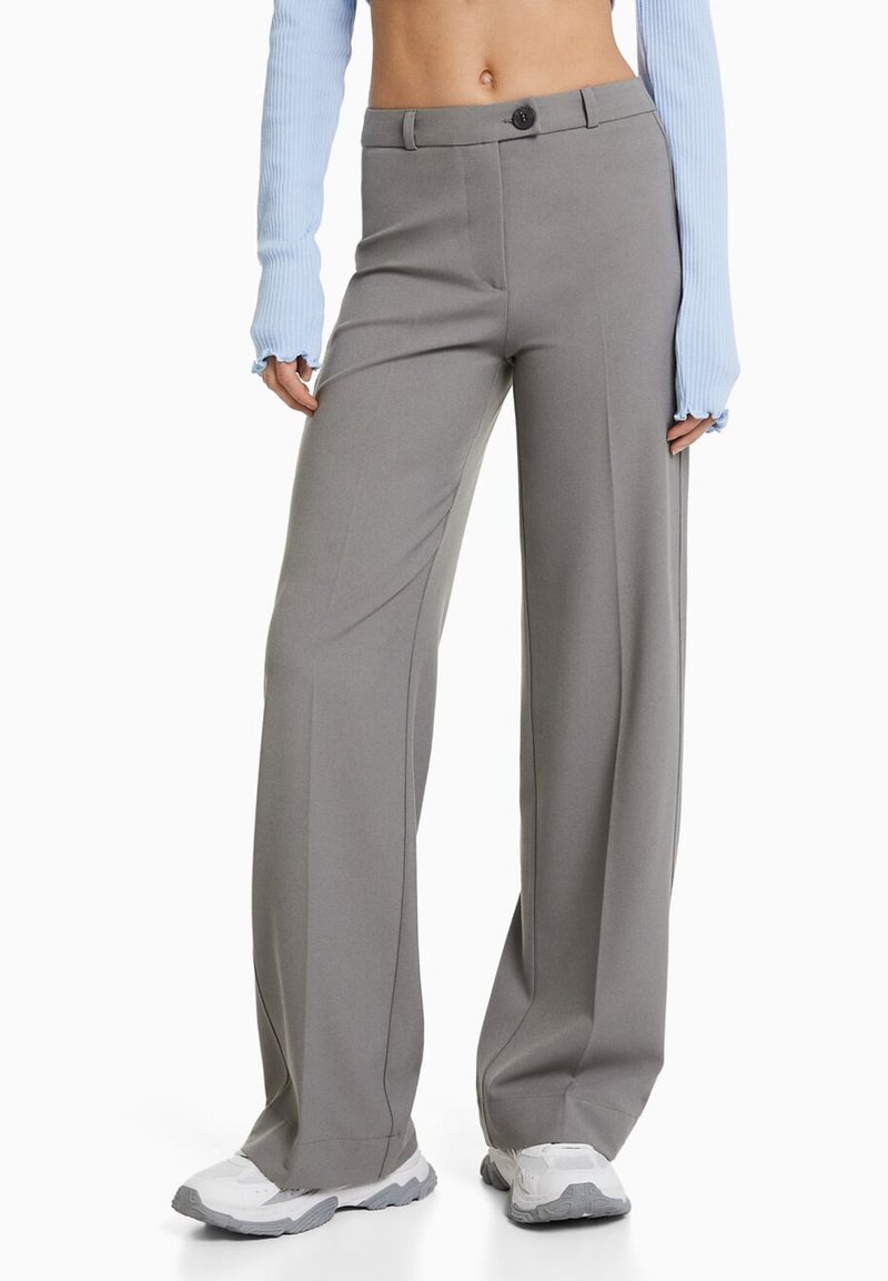Pantalons larges gris au toucher lisse, dotés d'une fermeture à bouton à l'avant et de plis subtils, associés à des chaussures de sport blanches.