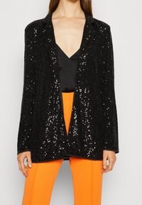 Blazer negro con lentejuelas y acabado texturizado, con delantero abierto y solapas con muesca, llevado sobre una blusa negra y pantalones naranjas.