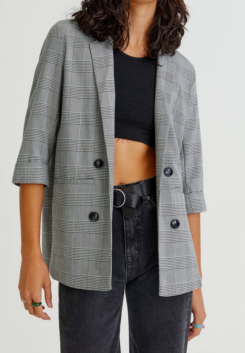 Femme portant un blazer à carreaux gris avec des manches trois-quarts, un top noir court, un jean noir taille haute et une ceinture avec boucle ronde.