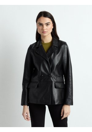 Kvinde med langt mørkt hår iført en sort læderdobbeltradet jakke over en olivengrøn top, stående mod en ensartet lys baggrund.