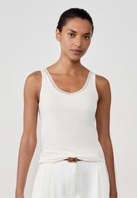 Donna con capelli scuri, indossa un top bianco senza maniche con bordo in pizzo, pantaloni bianchi e una cintura marrone, in piedi contro uno sfondo chiaro e uniforme.