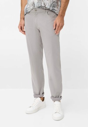 Mand iført lysegrå bukser med opkrogede ben, hvide sneakers og en lysefarvet skjorte med bladmønster, stående mod en hvid baggrund.