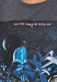 Grafische t-shirt met een blauw Darth Vader ontwerp tegen een sterrenachtergrond. Tekst "MAY THE FORCE BE WITH YOU" in het roze boven het beeld.