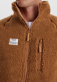 Brun fleecejakke med høj krave, to frontlynlåse og et stofmærke på venstre side. Tekstureret, blødt materiale med et fluffy udseende.