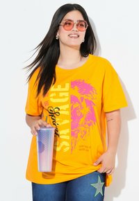 Camiseta naranja brillante con texto gráfico en negro y rosa, combinada con jeans azules que tienen un parche de estrella verde. Sosteniendo un vaso transparente y texturizado.