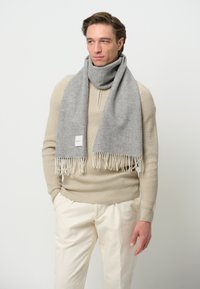 Grå uldscarf med frynse-detaljer, draperet om halsen, parret med en cremet striktrøje og beige bukser. Simpel, blød tekstur.