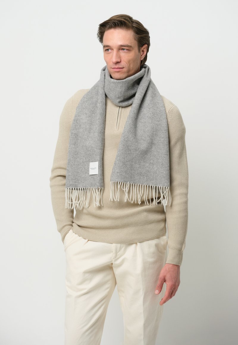 Grå uldscarf med frynse-detaljer, draperet om halsen, parret med en cremet striktrøje og beige bukser. Simpel, blød tekstur.