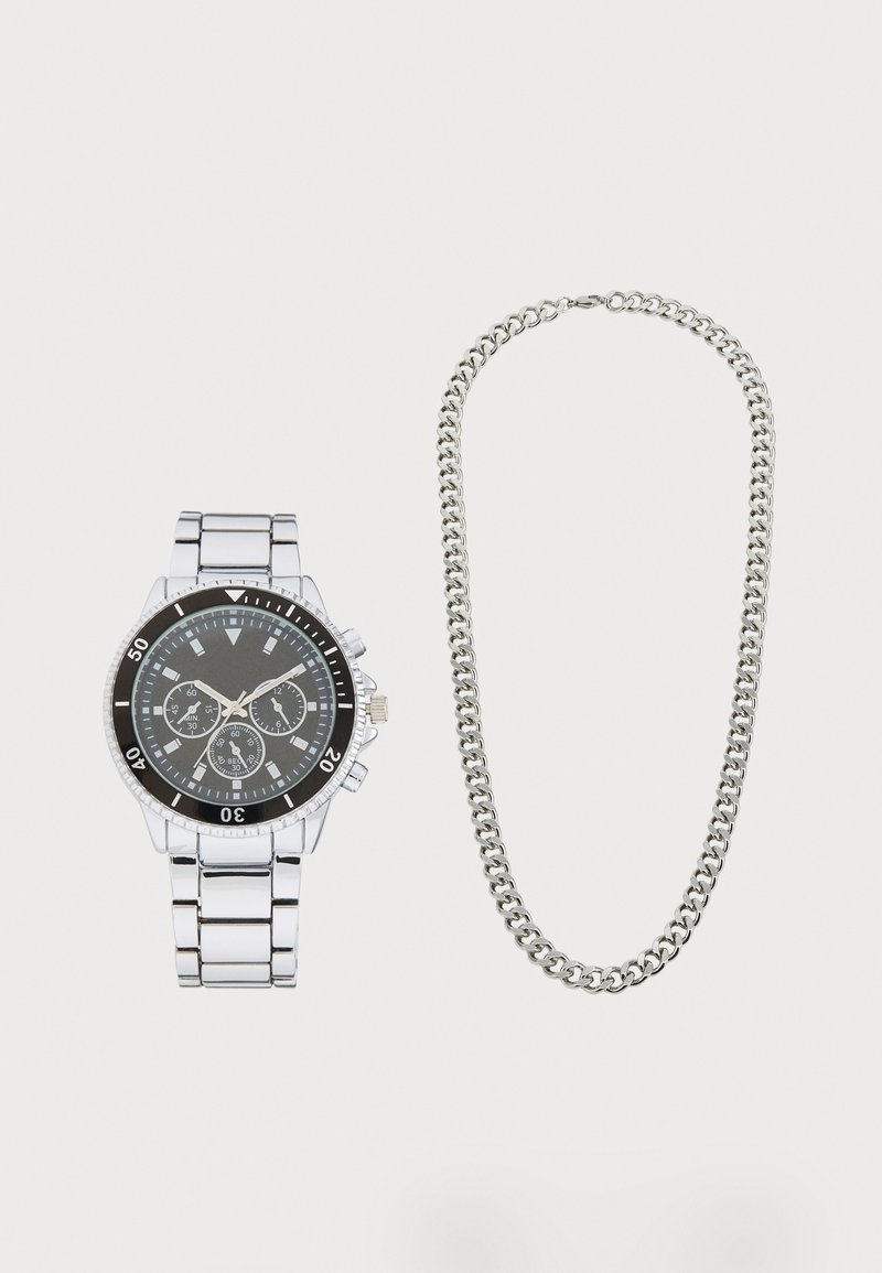 Ασημένιο μεταλλικό ρολόι χειρός με μαύρο κυκλικό καντράν και τρία υποκαντράν, δίπλα σε μια παχιά ασημένια αλυσίδα κολιέ με διασυνδεδεμένους κρίκους.