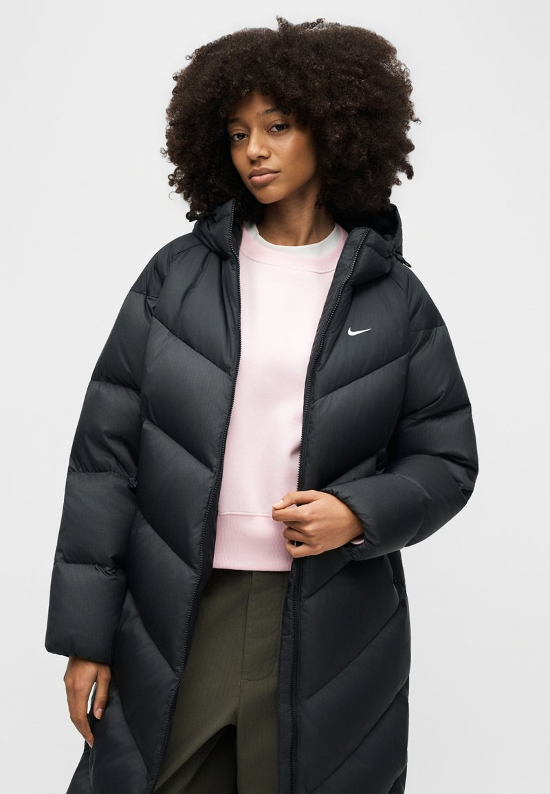 Frau mit lockigem Haar, die eine lange schwarze Steppjacke über einem hellrosa Sweatshirt und olivgrünen Hosen trägt, vor einem schlichten Hintergrund stehend.