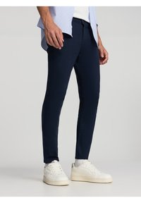 Pantaloni slim-fit blu navy in un tessuto morbido, con zip e chiusura a bottoni. Abbinati a sneakers bianche e una camicia azzurra chiara.