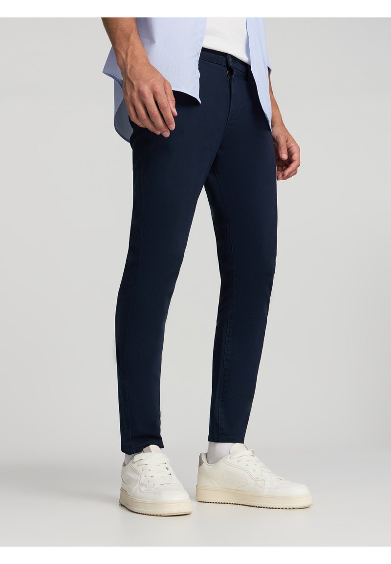 Pantaloni slim-fit blu navy in un tessuto morbido, con zip e chiusura a bottoni. Abbinati a sneakers bianche e una camicia azzurra chiara.