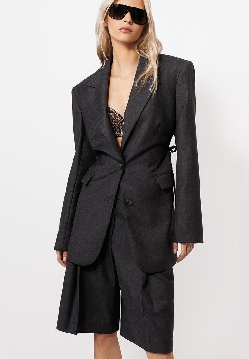 Szőke nő, aki túlméretezett sötétszürke blézert visel, hozzá illő térd alá érő rövidnadrággal, fekete mintás melltartóval és nagy fekete napszemüveggel.