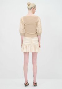 Top in maglia beige con maniche in pizzo gonfie, abbinato a una mini gonna crema con volant e arricciature. Visto da dietro, sfondo minimalista.