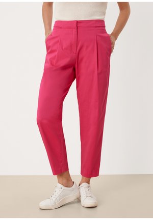 Person iført højtaljede, lyserøde skrå bukser med sidelommer og hvide sneakers, stående mod en ensfarvet baggrund.