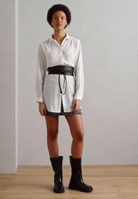 Witte blouse met knopen, zwarte tailleband en een gemusterde minirok met een rafelige zoom. Zwarte kniehoge laarzen maken de outfit compleet.
