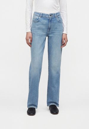 Personne portant un jean droit bleu clair, un haut à manches longues blanc et des chaussures noires, debout devant un fond uni.