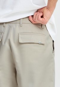 Beige bukser med glatt tekstur, med en baklomme med klaff og merke. Kombinert med en hvit t-skjorte, holdt av en hånd.