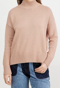 Hellrosa, gerippter Rollkragenpullover mit lässiger Passform und strukturiertem Strickmuster, kombiniert mit dunkelblauen Jeans.