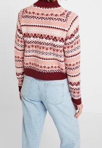 Maglione lavorato a maglia con un motivo bordeaux e rosa caratterizzato da design geometrici. Collo e polsini a costine, vestibilità corta, abbinato a jeans azzurri chiari.