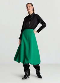 Schwarzes Hemd mit Knopfleiste, kombiniert mit einem lebhaften grünen Rock in Knielänge. Das Modell trägt glänzende, schwarze kniehohe Stiefel. Glatt fließender Stoff.