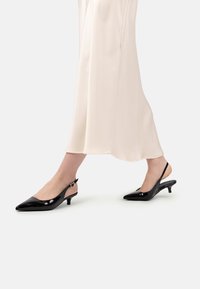 Femeie îmbrăcată cu o fustă din satin crem și pantofi slingback din piele lacuită negri, mergând pe o podea albă.