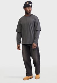 Tricou oversized gri închis stratificat peste o cămașă cu mânecă lungă, purtat cu blugi negri largi și saboți bej; o căciulă tricou neagră accentuată cu emagisment.