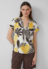 Blouse à manches courtes présentant un fond crème avec de grands motifs de feuilles jaunes et marron foncé, un décolleté en V et une coupe décontractée.