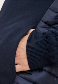 Granatowy element odzieży łączący gładką dzianinę z pikowanym, puchowym materiałem. Posiada ściągacz na mankiecie oraz rękę wsuniętą w odzież.