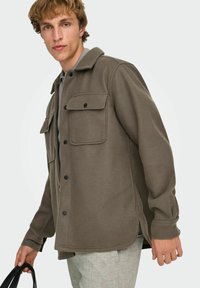Veste en polaire marron avec un col à revers, deux poches poitrine et des boutons-pression. Le matériau est doux avec une finition légèrement texturée.