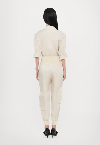 Helles beiges Jumpsuit mit kurzen Puffärmeln, elastischem Taillenbund und Cargo-Taschen; kombiniert mit schwarzen High Heels.