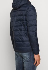 Navy puffer jacket with a hood, featuring quilted design, smooth texture, and elastic cuffs, worn over light blue jeans.