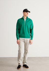 Zielony sweter z suwakiem przy szyi i prążkowanymi wykończeniami, w połączeniu z jasnoszarymi dżinsami i czarnymi trampkami, na neutralnym tle.