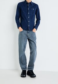Camisa de mezclilla oscura con botones y bolsillo en el pecho, combinada con jeans gris desgastados y zapatillas negras. Diseño sencillo, ajuste casual, mínimo detalle.
