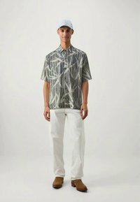 Camicia a maniche corte, fantasia nei toni del verde e beige, con una vestibilità ampia, abbinata a pantaloni bianchi e scarpe in suede marroni.
