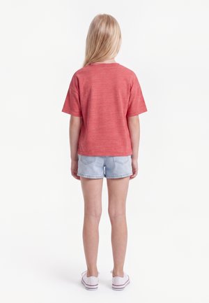 Enfant aux cheveux blonds et raides, de dos, portant un t-shirt rouge à manches courtes, un short en jean clair et des baskets blanches sur fond blanc.