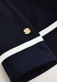 Marineblauer Stoff mit weißem Streifendetail und kleinem rundem goldfarbenem Knopf, graviert mit dem Buchstaben B, auf den Stoff genäht.