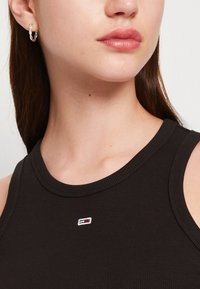 Fekete bordázott ujjatlan felső kerek nyakkivágással és kicsi hímzett logóval. Ezüst karika fülbevaló látható, sima bőr textúrával.