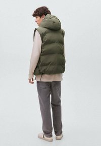 Olivnozeleni zimski prsluk s kapuco, nošen preko bež majice. Uparen s sivoj trapericama i svijetlim tenisicama. Jednostavan dizajn, bez vidljivih uzoraka.