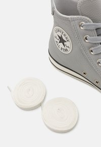 Adidași gri din piele, de tip high-top, cu logo-ul unei stele alb-negre, având ochiuri metalice, șireturi gri și două șireturi albe spiralate.