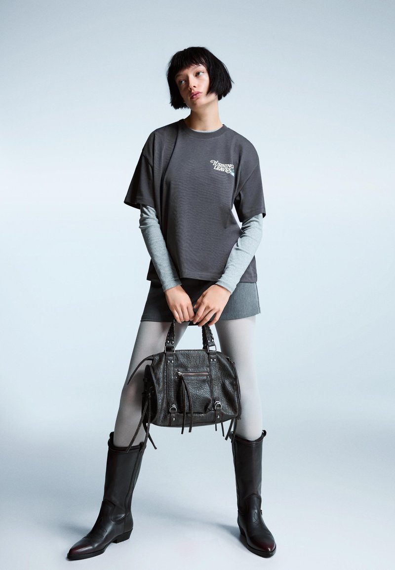 Tricou oversized gri, purtat peste o bluză cu mânecă lungă gri deschis, asortat cu o fustă gri și colanți gri deschis. Bocanci negri până la genunchi și o geantă neagră texturată.