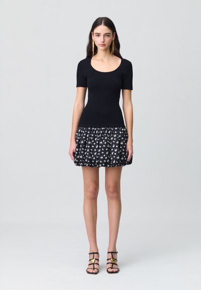 Haut noir ajusté à manches courtes avec un imprimé floral, jupe plissée en noir et blanc. Sandales noires à lanières avec des accents dorés.